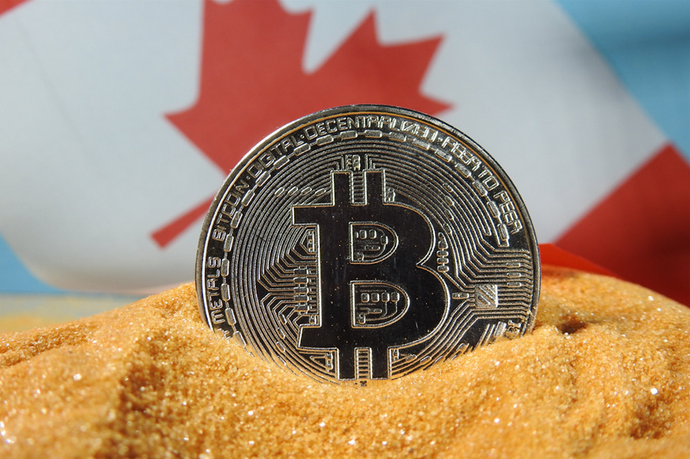 A silver Bitcoin in orange sand with a Canadian flag in the background (Source: unsplash)